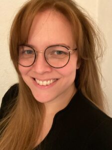 Ein Bild von Rebekah Schmidt. Eine lächelnde Frau mit langen, rotblonden Haaren und einer schwarz umrandeten Brille,